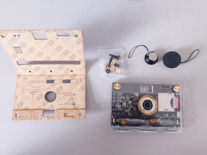 A top down shot of everything spread on a desk. On the left is an unfolded cork case. On the right the bottom half is taken up by a Paper Shoot in a Croz acrylic case. Above the cam is a small bag of screws, macro lens attachment and wide angle lens attachment.