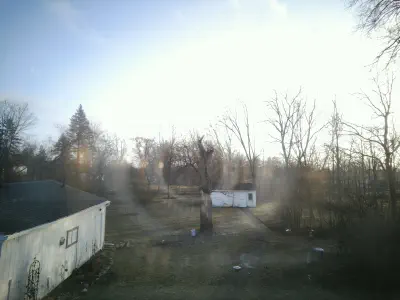 (In color) An elevated shot of a backyard. On the left is a simple 1 story garage, in the center is a willow tree and further back is a white shed. On the right is a lot of landscaping and critter feeders.