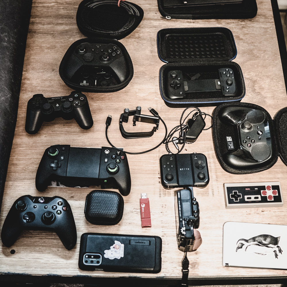 A top down shot of 8 controllers, 2 phones, a camera, and varied accessories. The full set of controllers are: Microsoft Elite Series 2, Sony PS4, PowerA MOGA XP-7, Xbox One, Kishi v2, 8bitdo NES, Jungle Cat, and Steam.