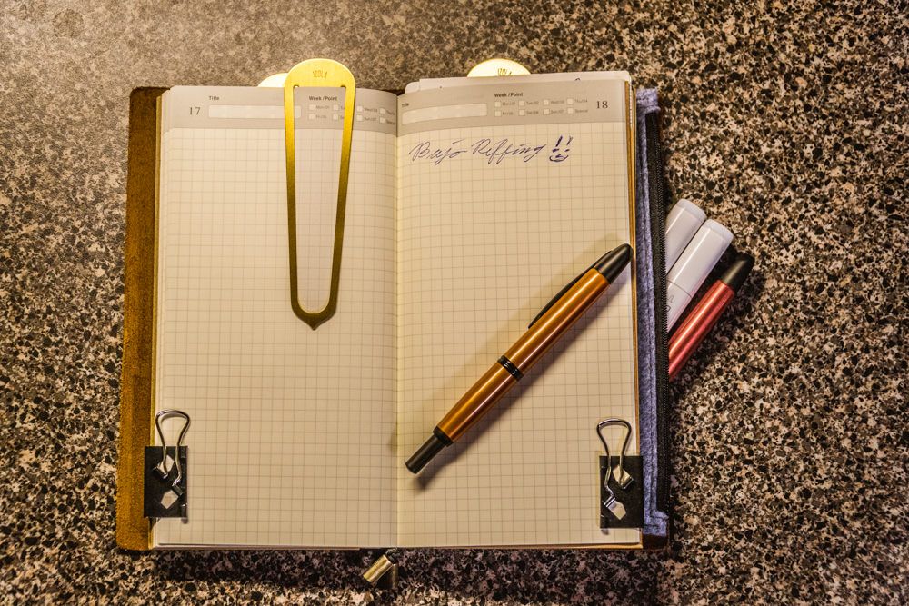 A wanderer's travel notebook open to the center with two fountain pens, two markers, two binder clips and a bookmark shown